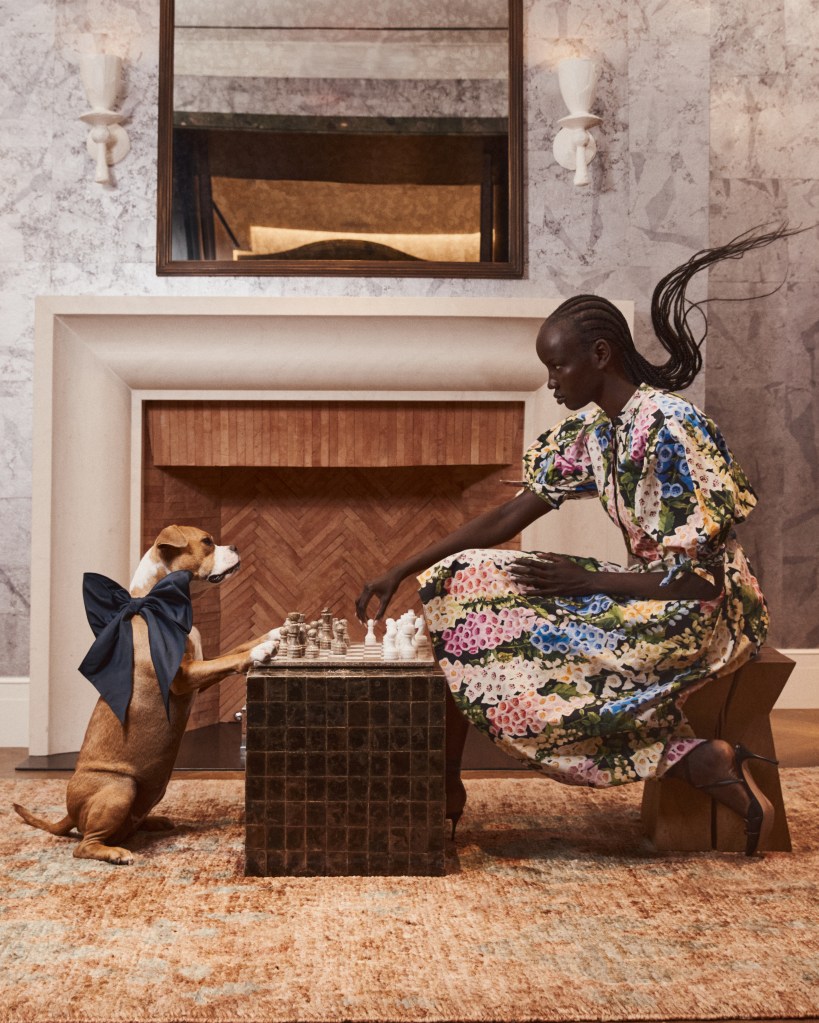 A Black woman in a floral dress plays chess with a brown dog wearing a blue bow.