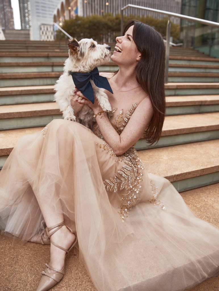 A woman in a gold Oscar de la Renta gown sits on steps, holding a white dog wearing a matching black bow tie from the Simon Sits collaboration.