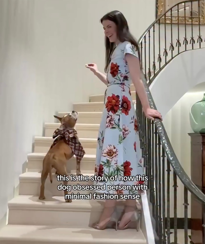 A woman in a floral dress and a dog in a plaid neckerchief stand on a white staircase with a black railing, looking at each other.
