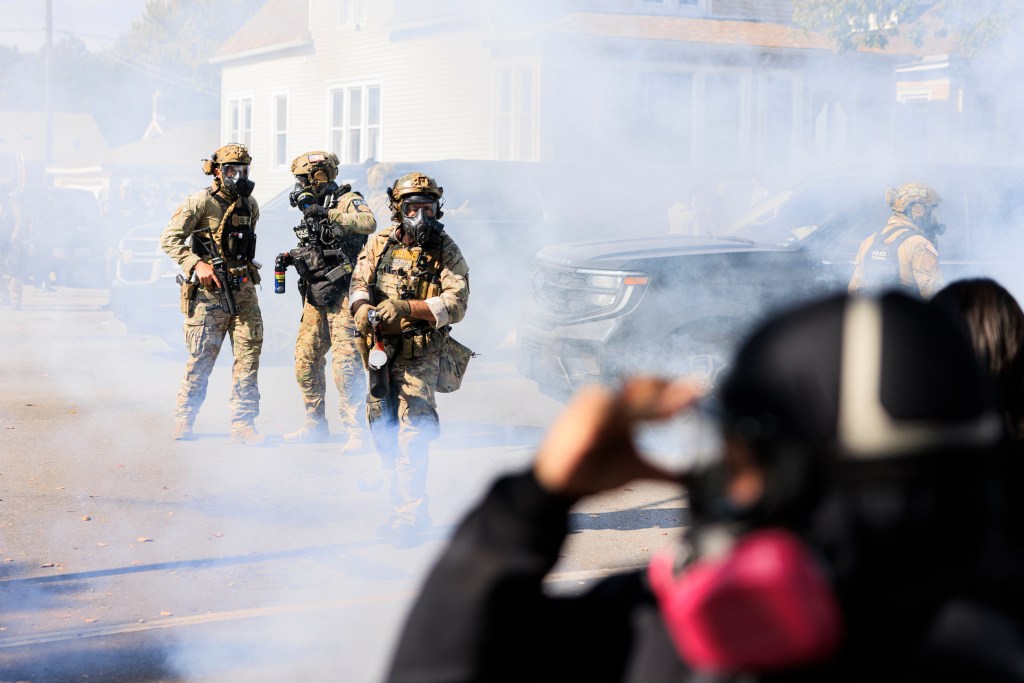 Government officials in tactical gear and gas masks amid smoke during a protest.