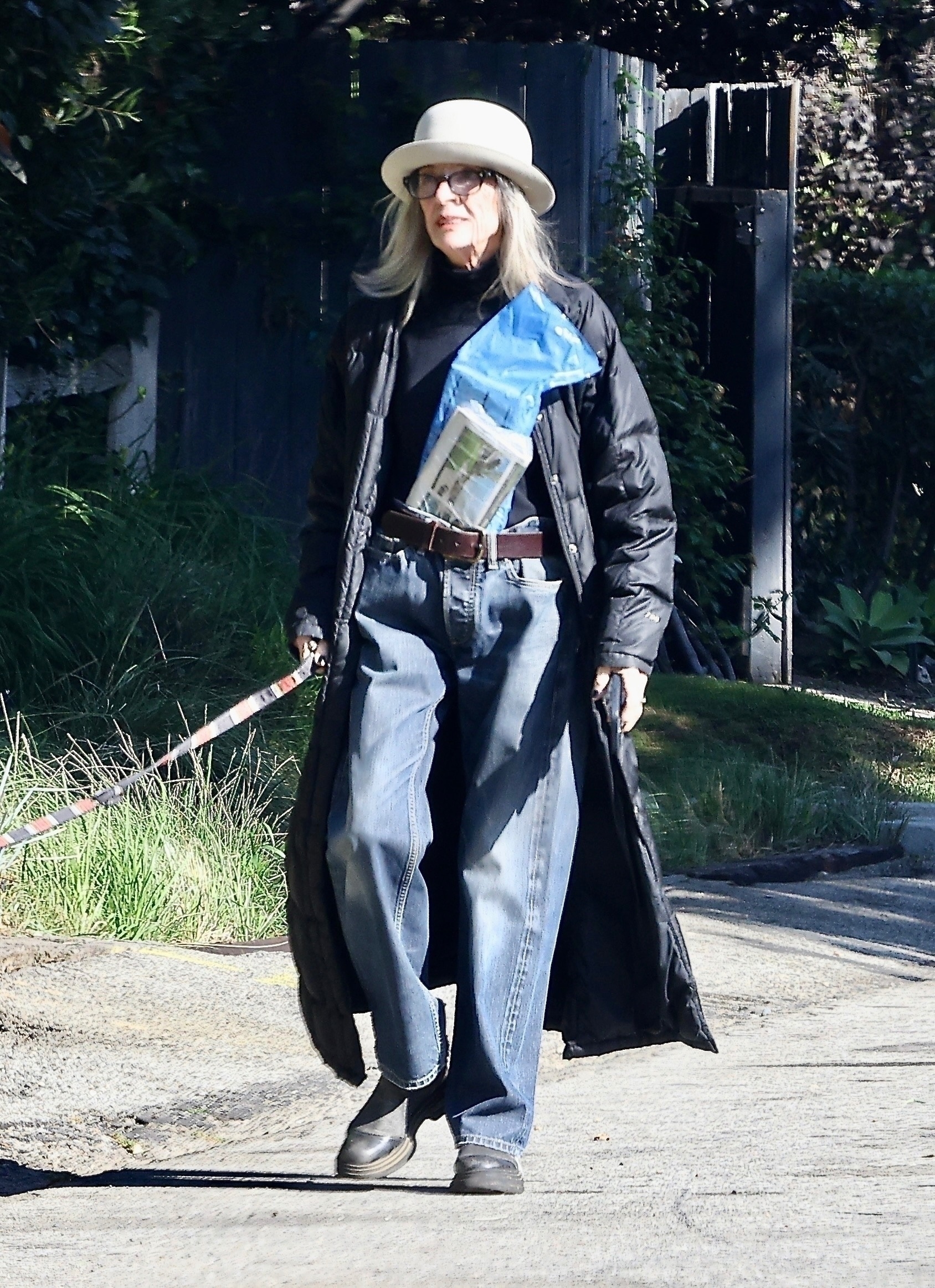 Diane Keaton wearing a black trench coat, black turtleneck, black pants, black boots, black glasses, and a gray hat, holding a face mask.