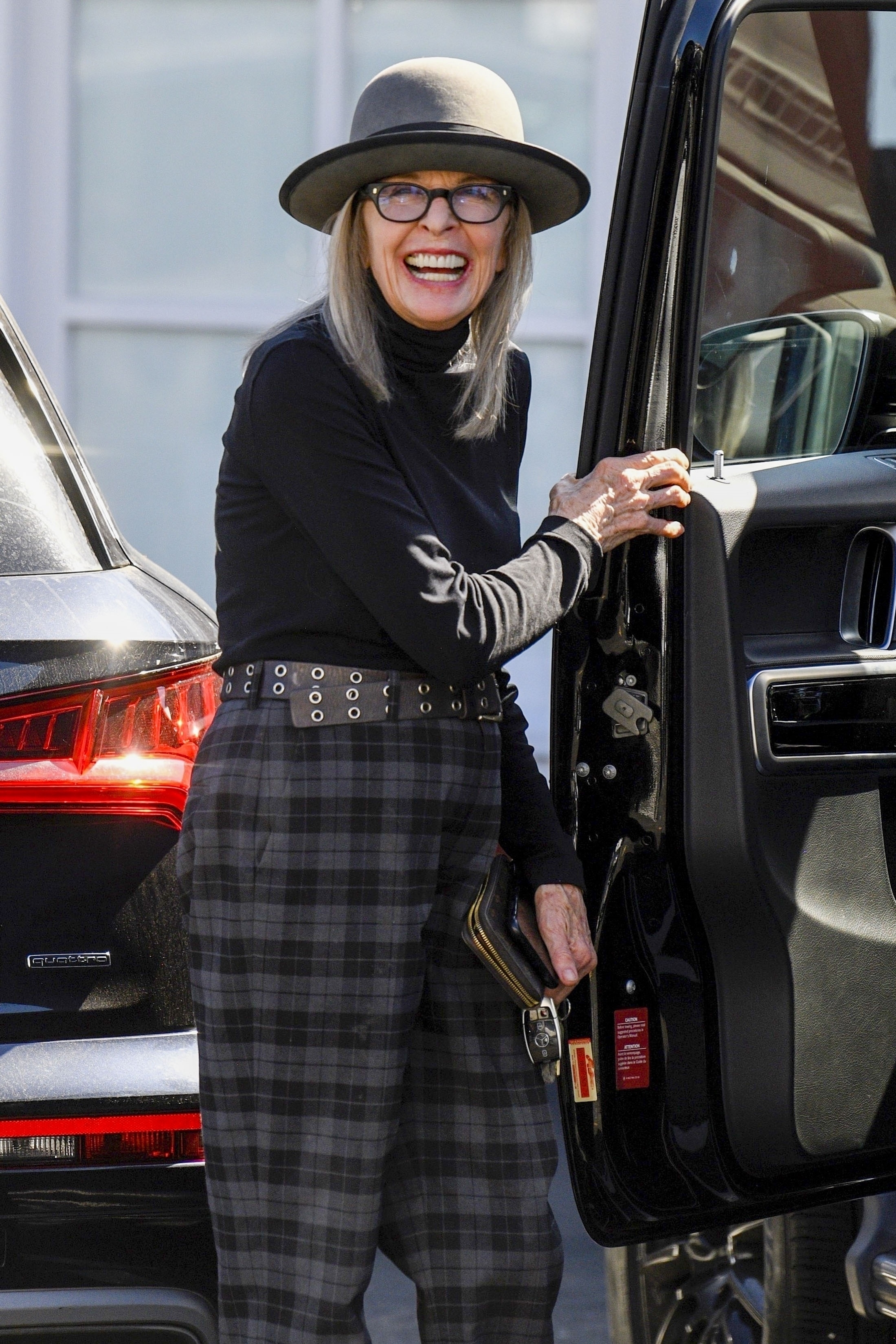 Diane Keaton smiling and wearing a hat, glasses, a black turtleneck, a studded belt, and plaid pants, while getting out of a black car.