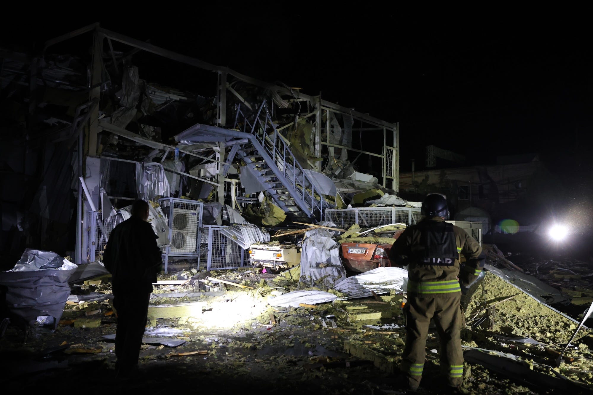 Two figures look upon twisted metal of a wrecked building at nighttime
