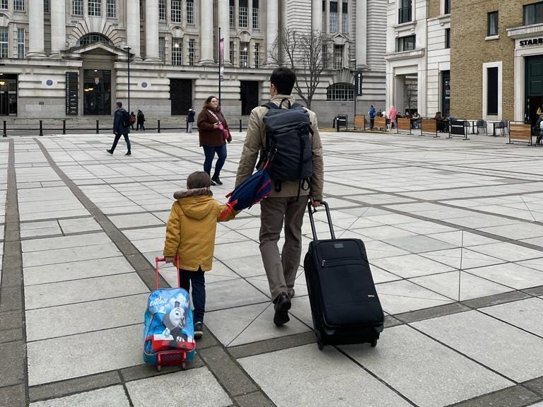 The writer walking with his son in London. They're both pulling suitcases and facing away from the camera.