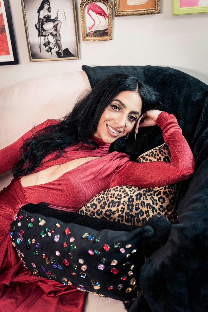Ester Yuabova lying on a couch with a black cushion and a leopard print cushion, wearing a red dress with a cutout, looking at the camera and smiling.