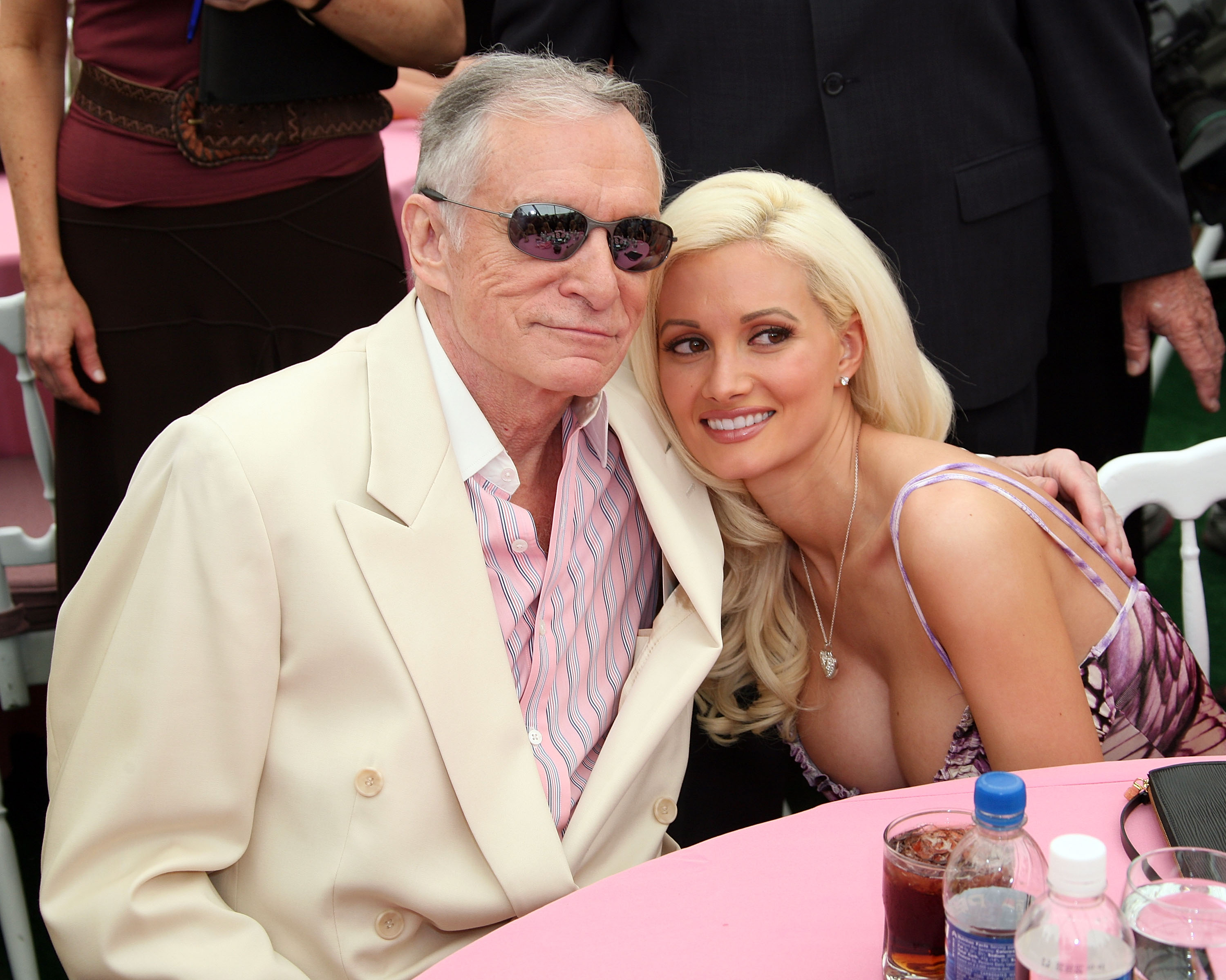 Hugh Hefner in a white suit and sunglasses, sitting next to Holly Madison, who wears a purple and white patterned top.