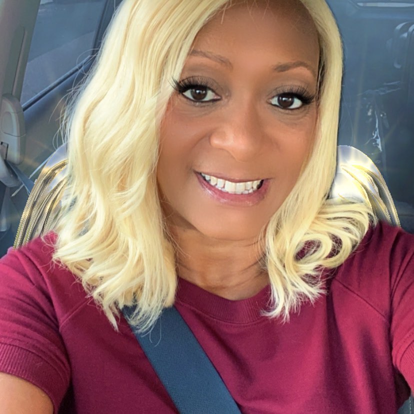 Woman with blonde hair and a maroon shirt smiling in a car.