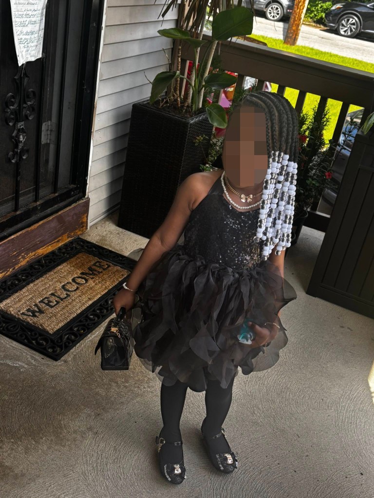 A young girl in a black dress with beads in her hair stands on a porch with a welcome mat.