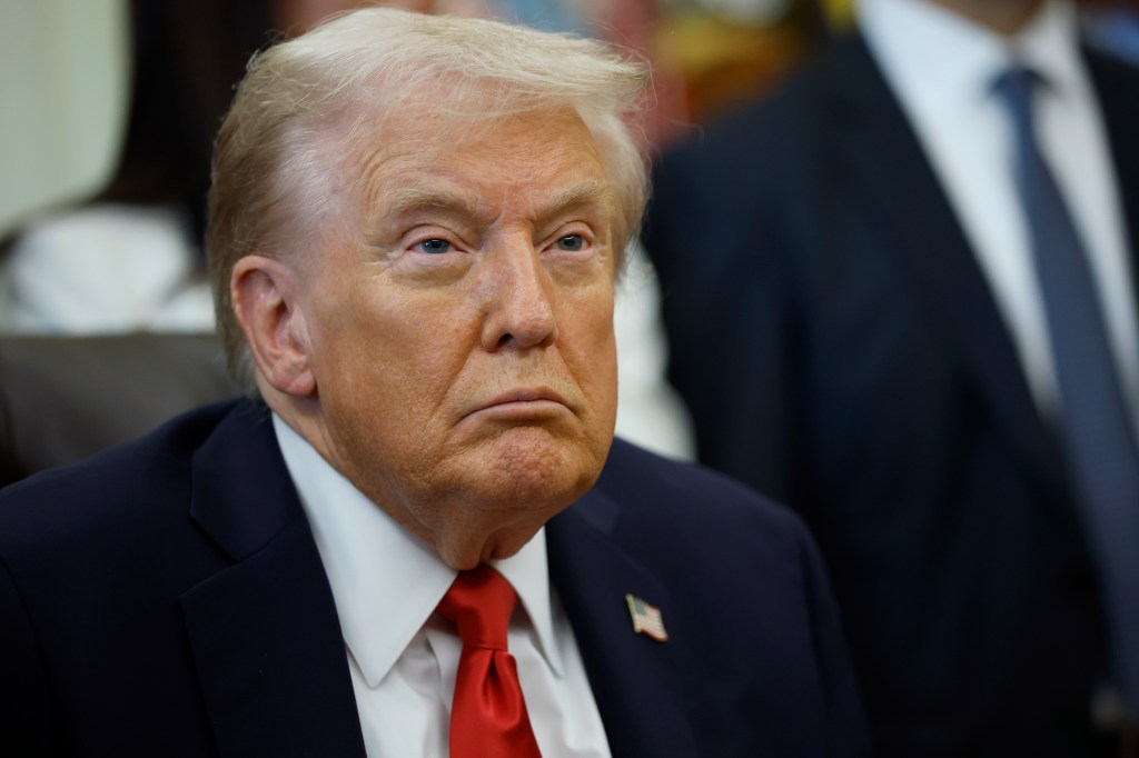Donald Trump in a suit and red tie listening to officials in the Oval Office.