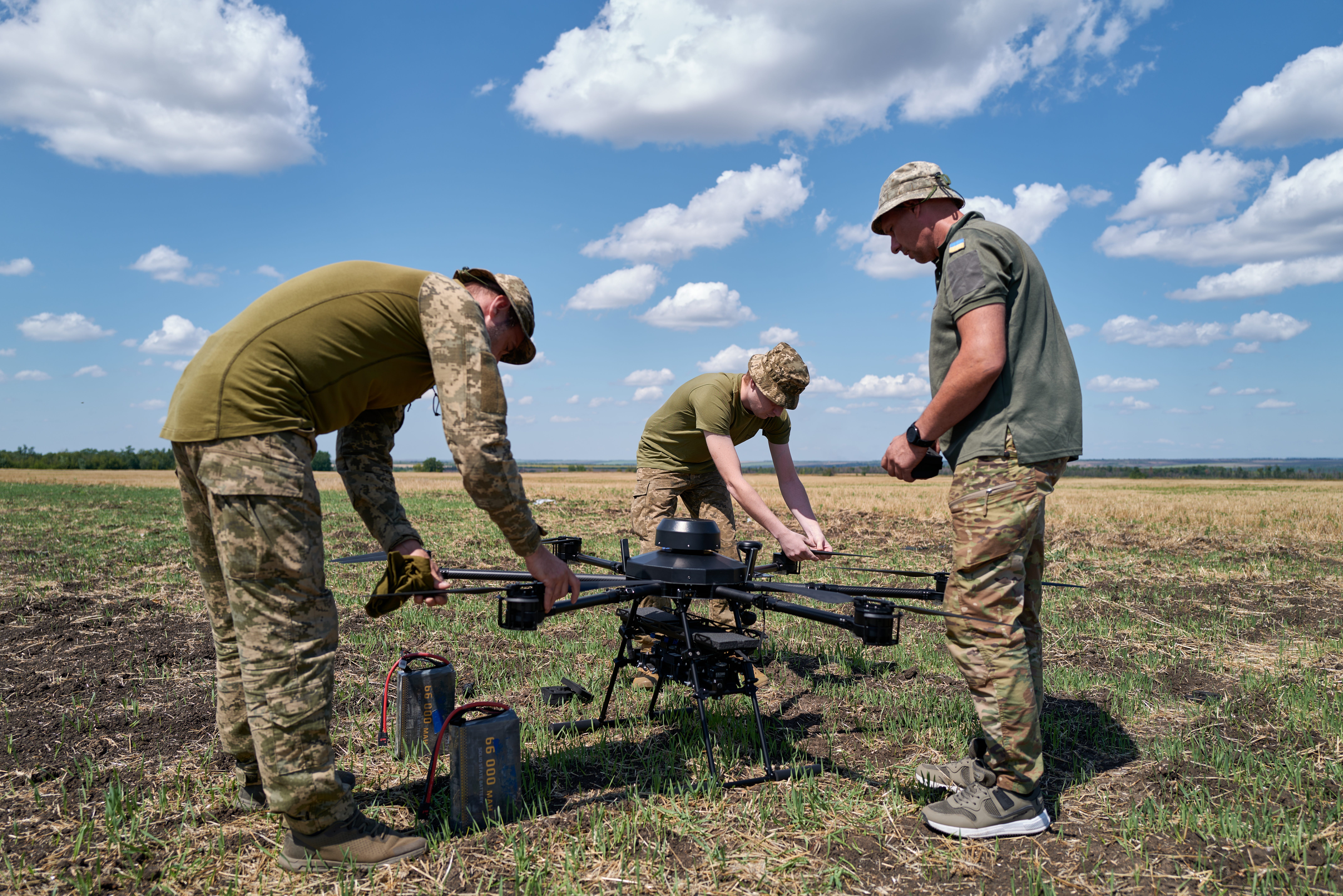 Three men in camoflage gear and green t-shirts stand around a drone in a field under a blue sky