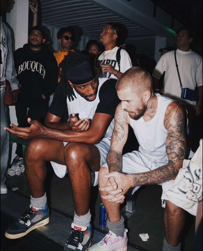 Justin Bieber in a white tank top and shorts, with heavily tattooed arms and legs, sitting and talking with another man.
