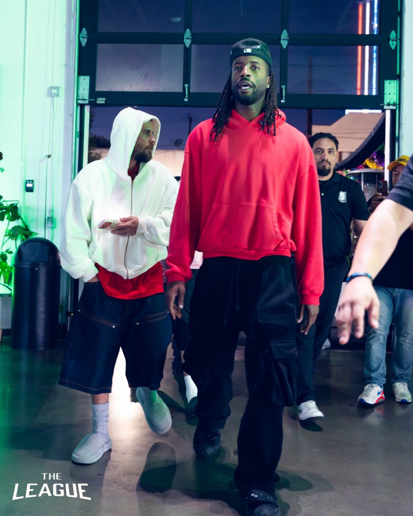 Justin Bieber with a beard and hooded shirt walking next to another man with long braids and a red hoodie.