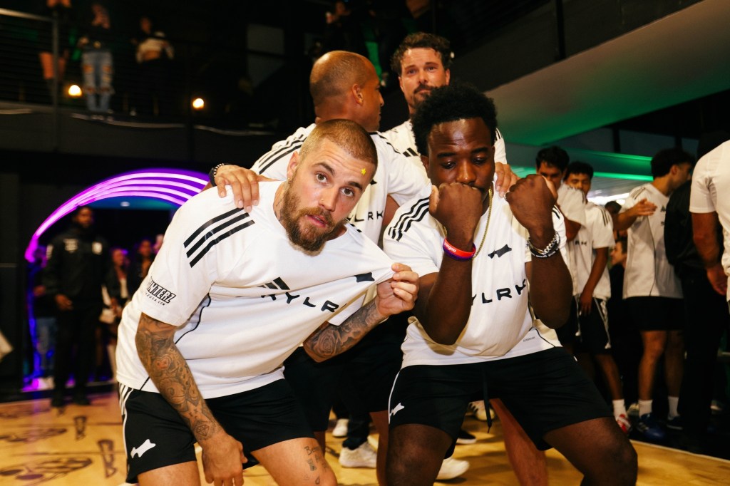 Justin Bieber poses with a man, surrounded by other men on a basketball court.
