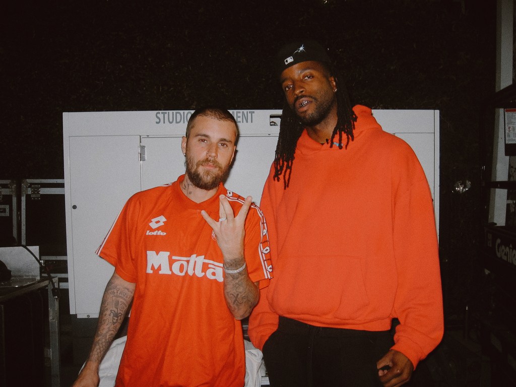 Justin Bieber standing next to another person with dreadlocks, both wearing orange shirts.