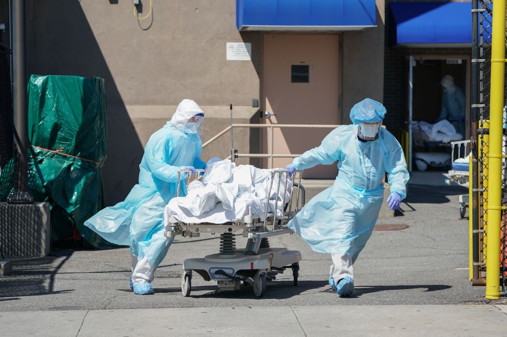 Medical workers in PPE transport a body on a gurney to a refrigeration truck outside a hospital.