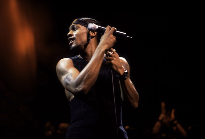 A color photograph of D'Angelo performing and holding the microphone stand on a darkly lit stage