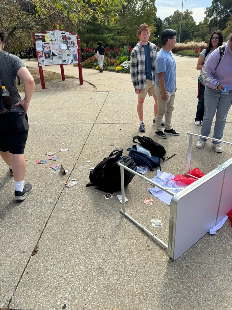 A table is overturned with items scattered on the ground. A group of people are standing around the mess.