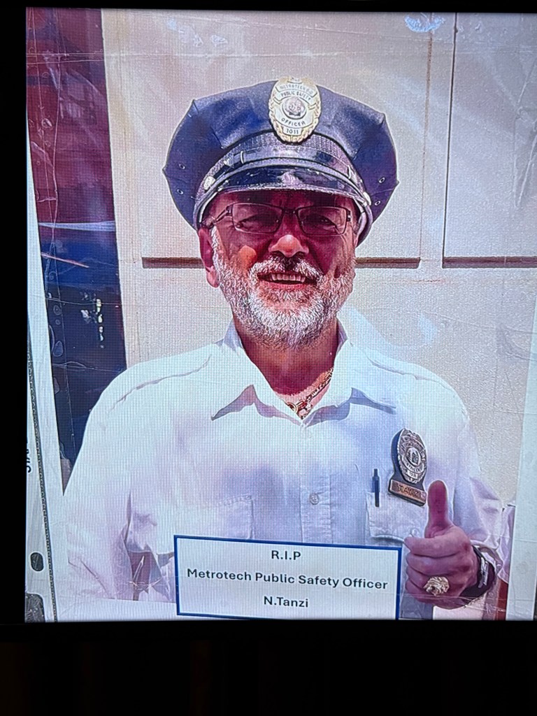A photo of security guard Nicola Tanzi shown on a TV screen, wearing a cap, glasses, and a badge, giving a thumbs-up.