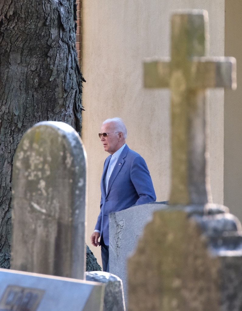 Joe Biden, wearing sunglasses and a blue suit, walking through a graveyard.