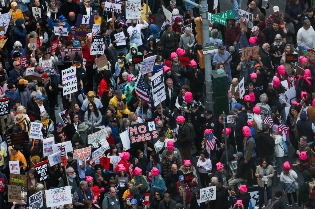 People protest in Times Square against Trump's policies, holding various signs including 