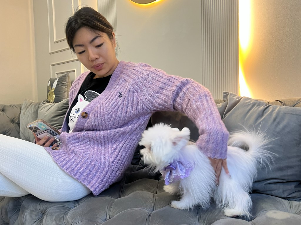 A woman in a purple cardigan and white pants sits on a gray couch, holding a phone and petting a small white dog with a purple collar.