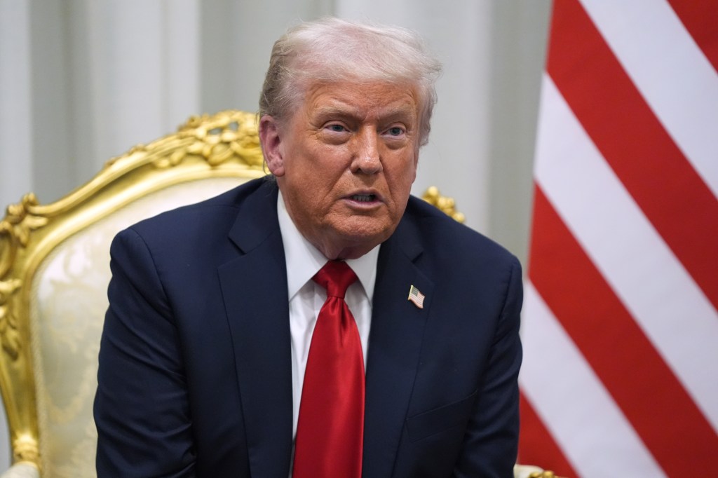 President Donald Trump speaking while seated in a gold-colored chair, with a red and white striped flag behind him.