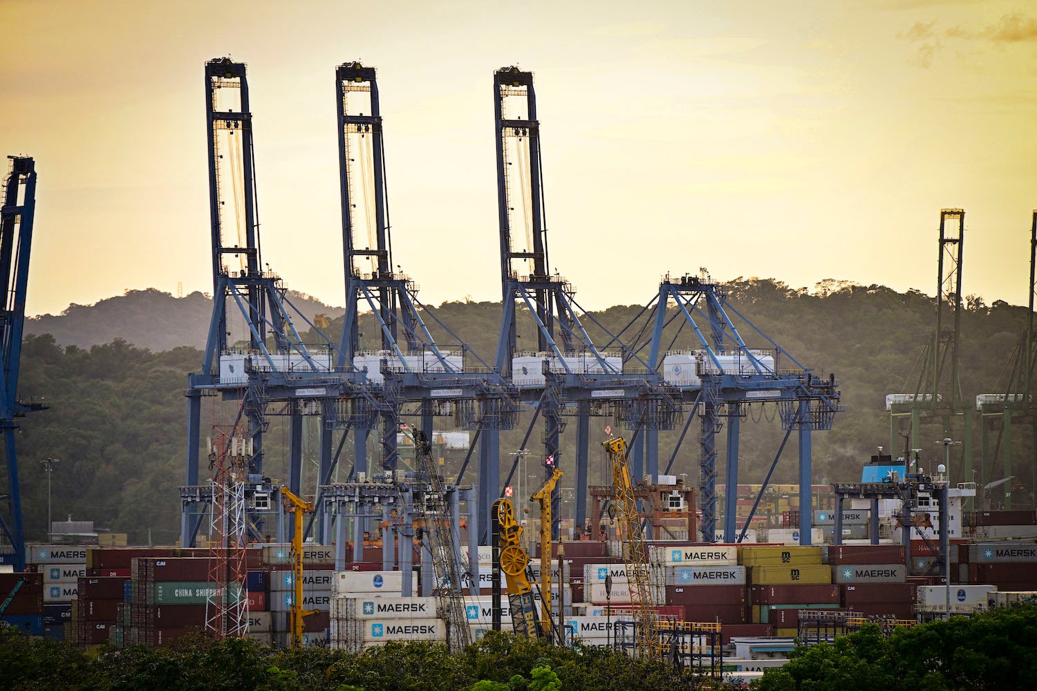Cranes extend into a yellow sky high above stacks of container ships at a port on the Panama Canal.