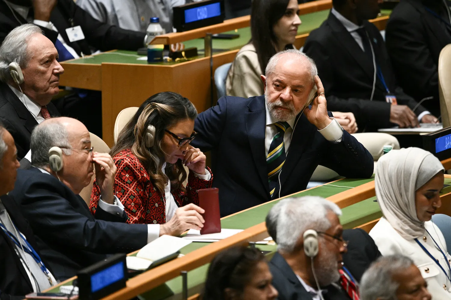 Brazilian President Luiz Inácio Lula da Silva leans on his left elbow while listening via an audio device on his left ear. He is wearing a striped tie and is seated at a green desk among other dignitaries.
