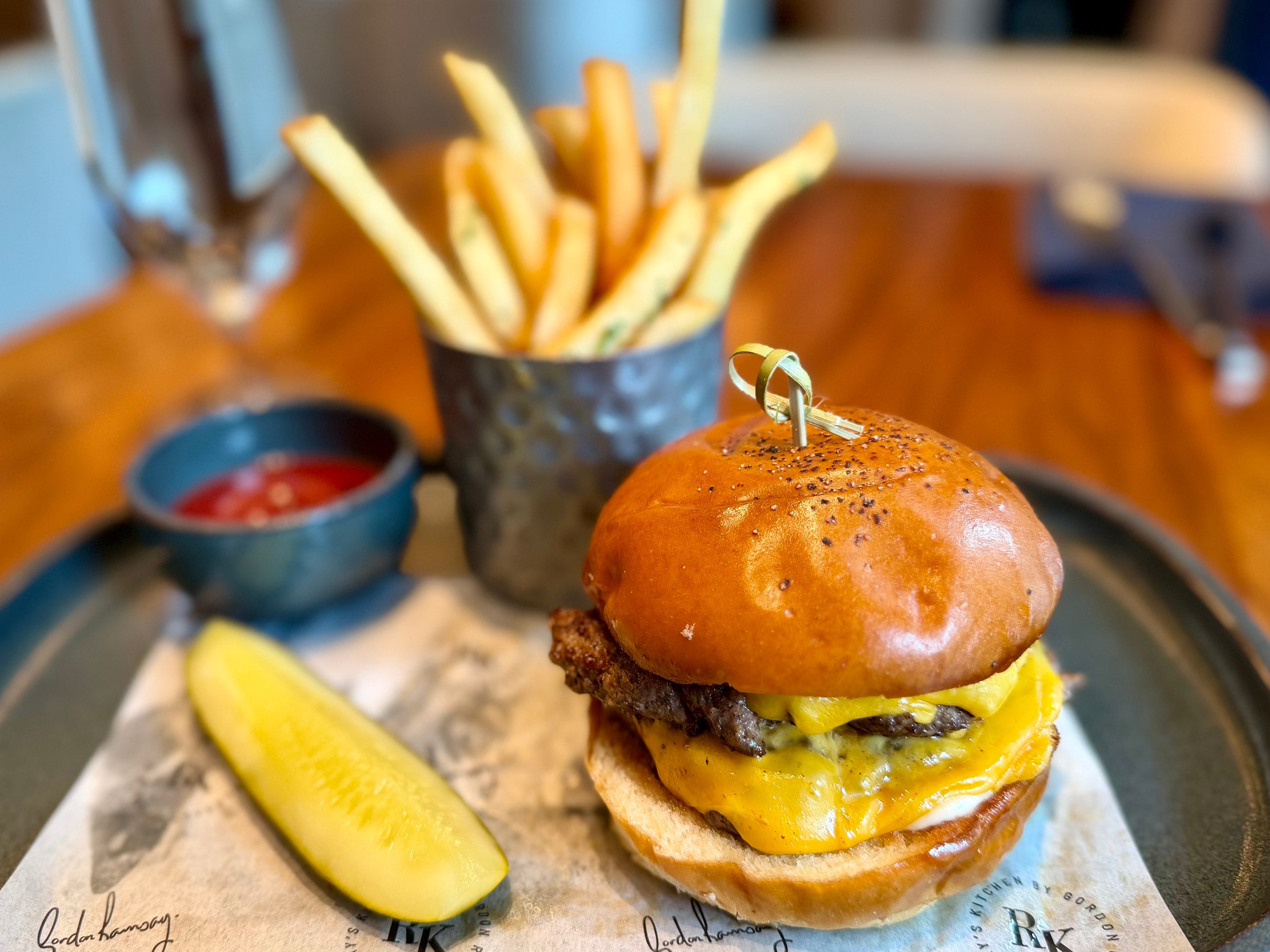 A plate with a burger, a cup of fries, and a side of ketchup, and a pickle on a plate at Ramsay's Kitchen.