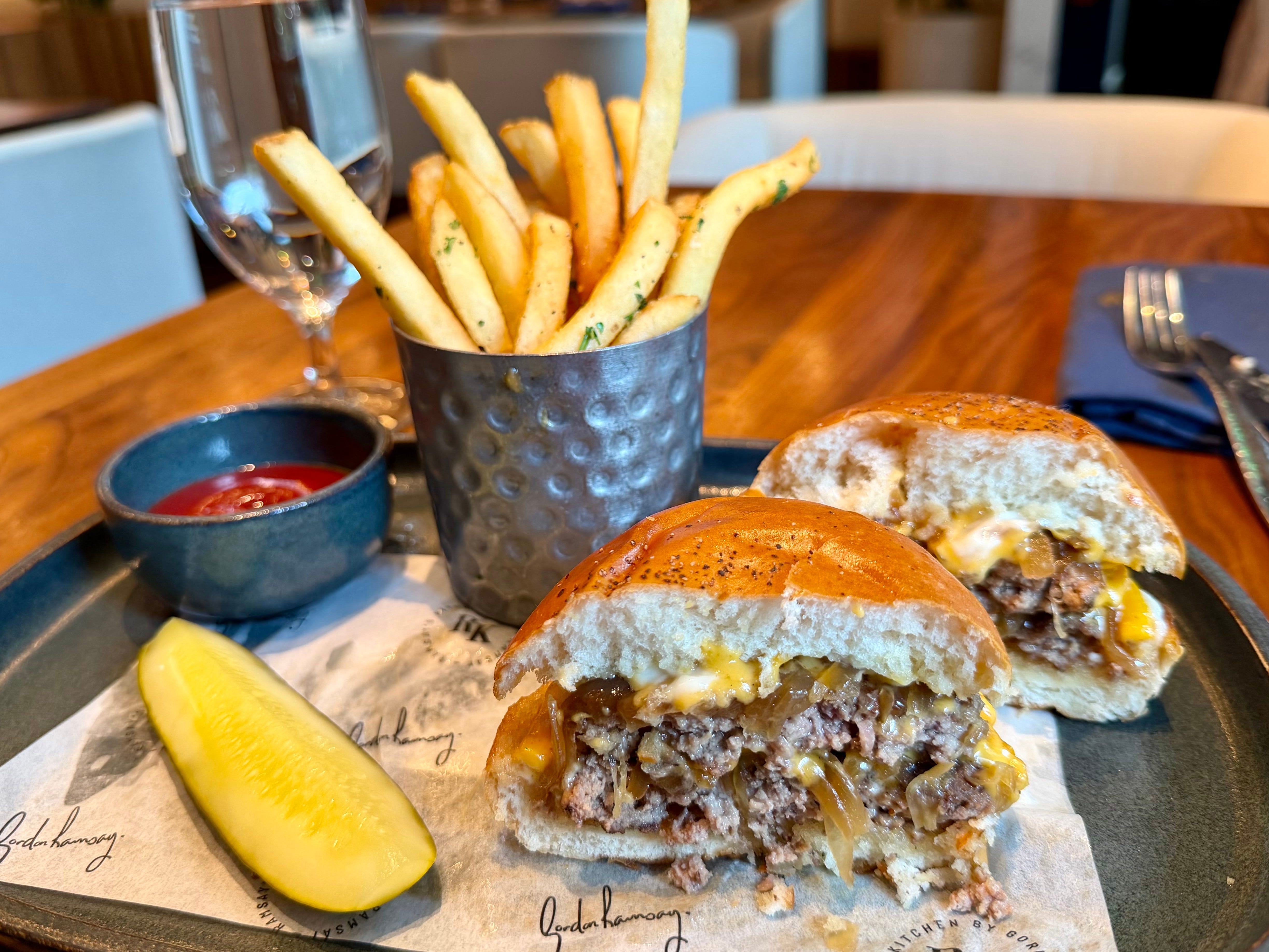 A burger cut open in half on a plate with a cup of fries, a pickle, and a side of ketchup.