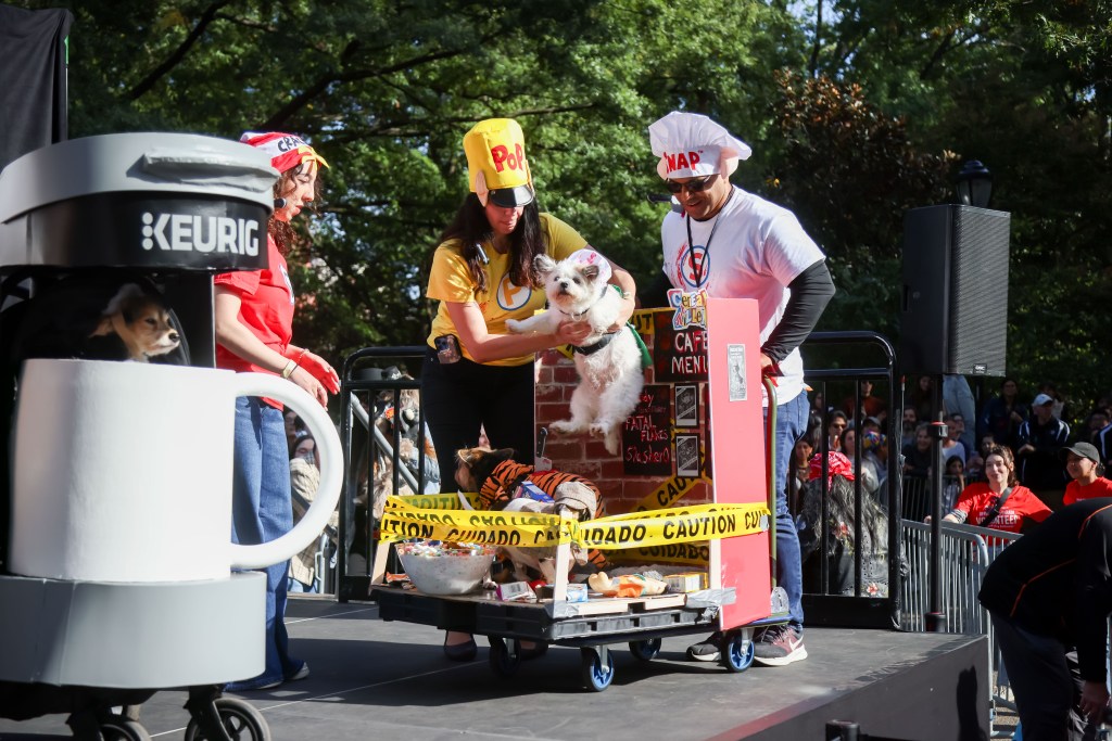 A person in a coffee costume, a woman in a popcorn costume holding a white dog, and a man in a chef's hat standing next to a cart with dogs in tiger costumes and a 