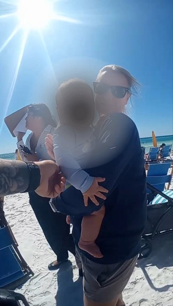 A woman in sunglasses holding a baby on a sunny beach, another person in uniform standing behind them.