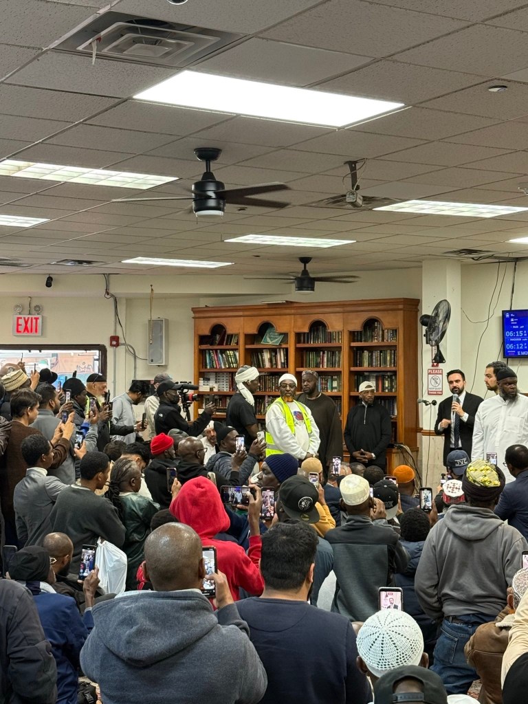 A crowd of men recording a speaker at a podium in a mosque with their phones.