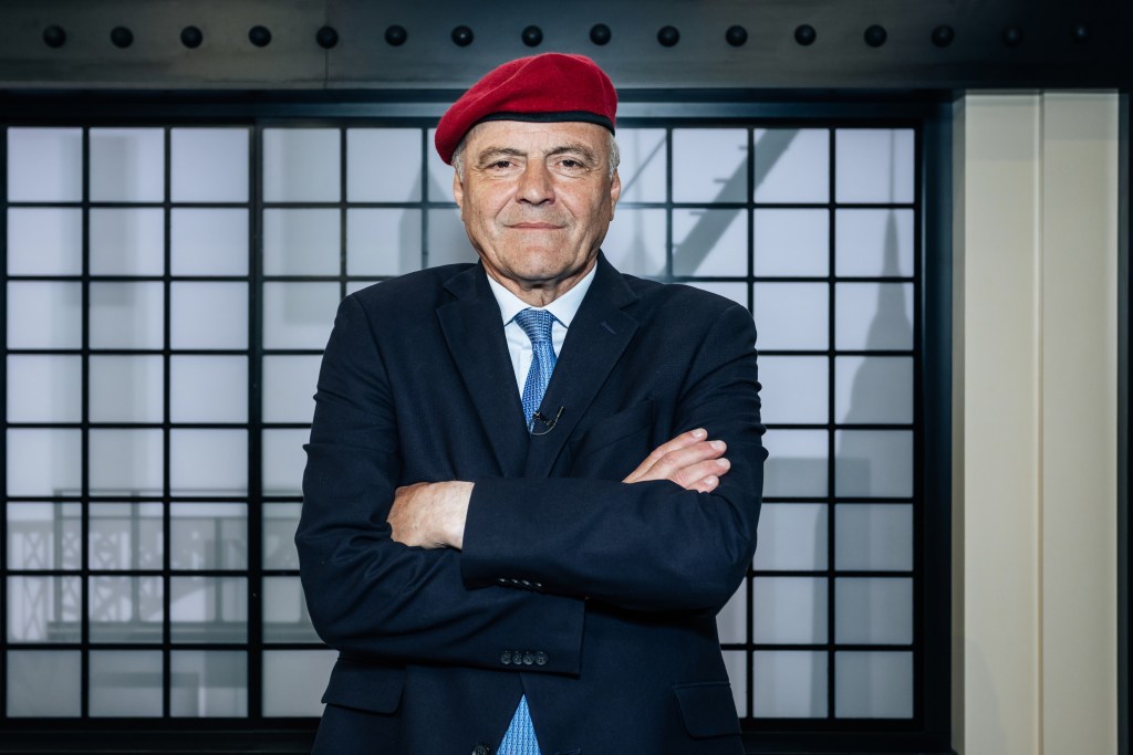 Curtis Sliwa in a dark suit and red beret, with his arms crossed, posing in a studio.