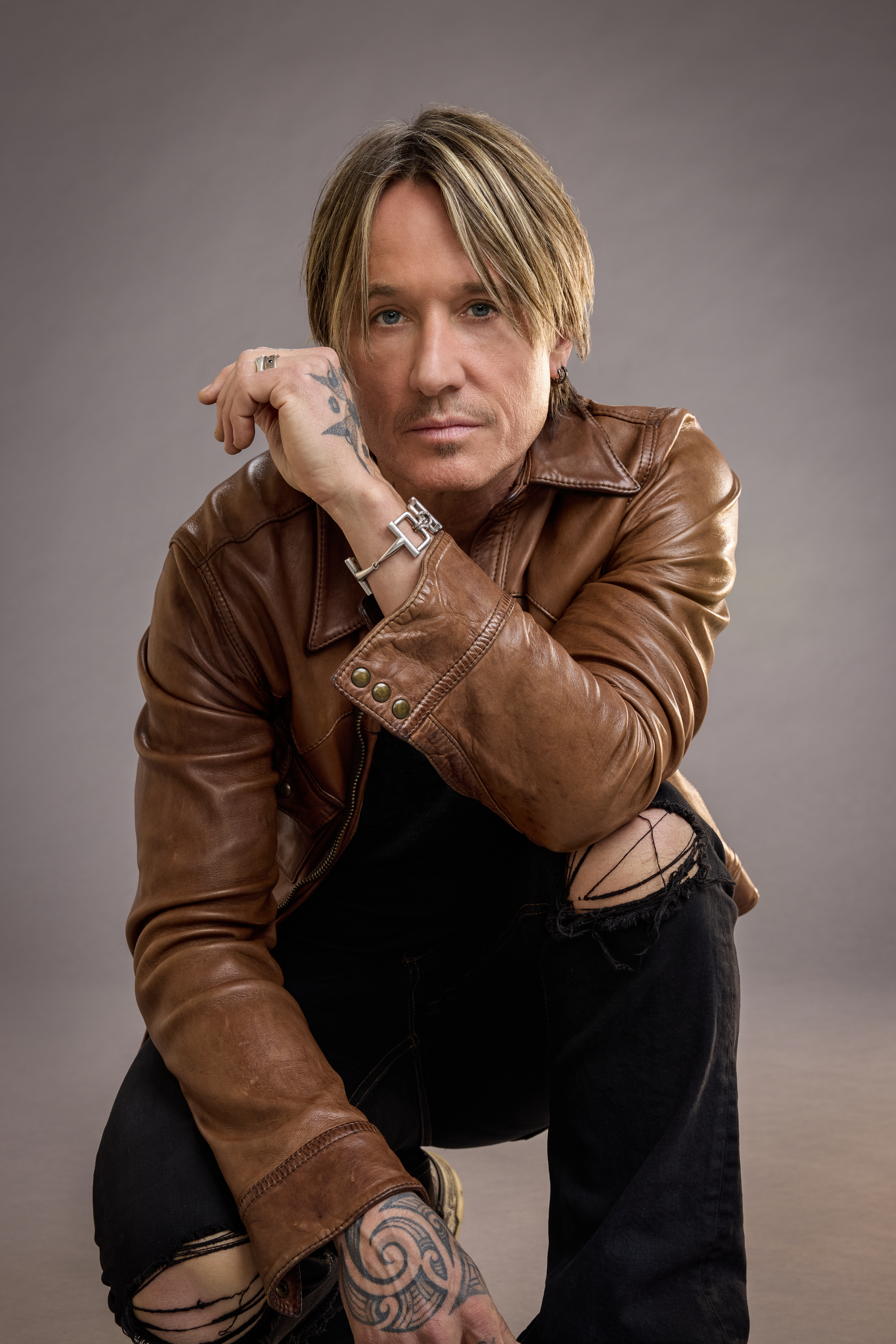 Keith Urban, Headliner and Executive Producer of THE ROAD, crouching, wearing a brown leather jacket and torn black jeans.