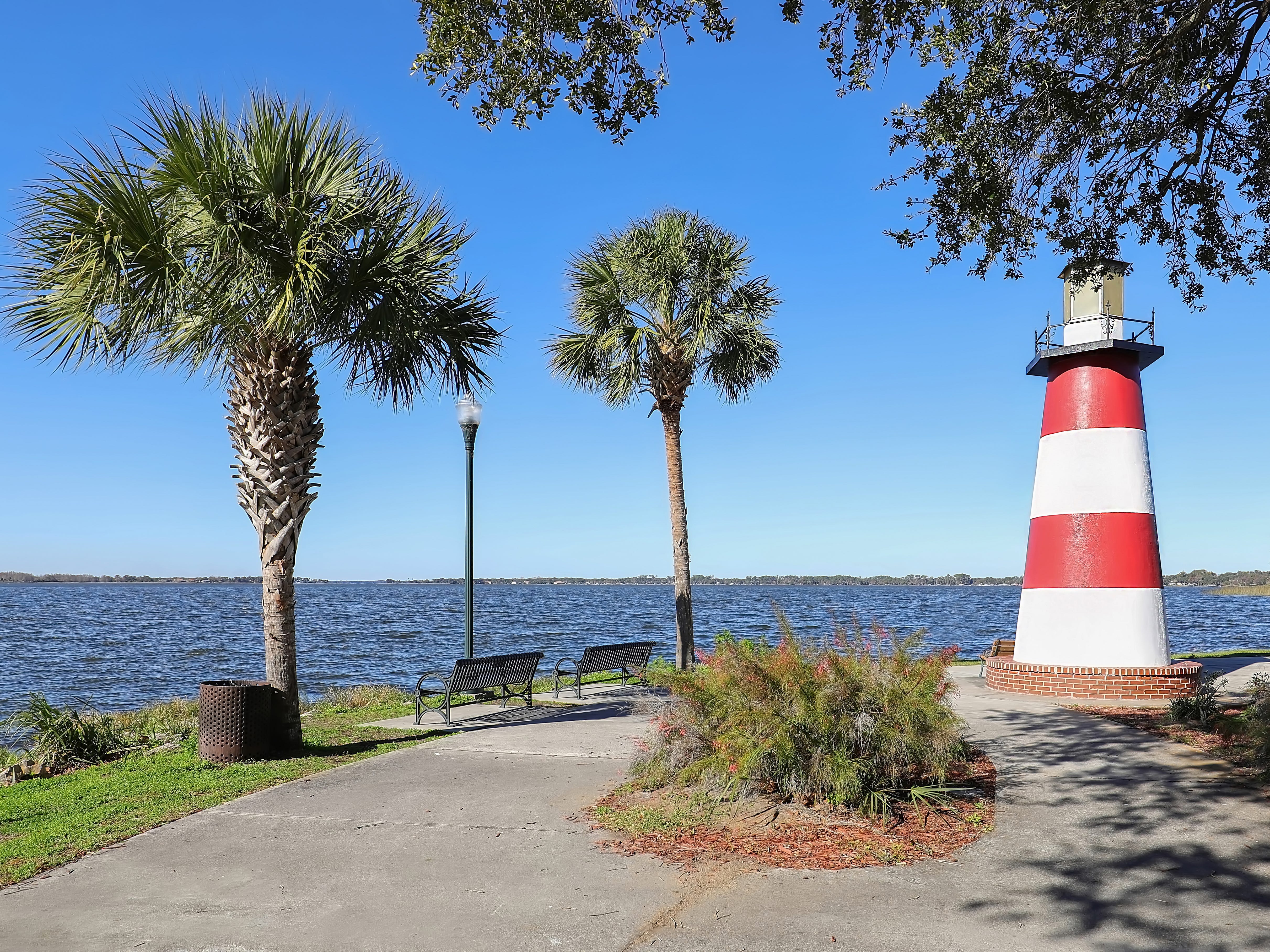 A small lighthouse near the water, with palm trees and benches surrounding it.