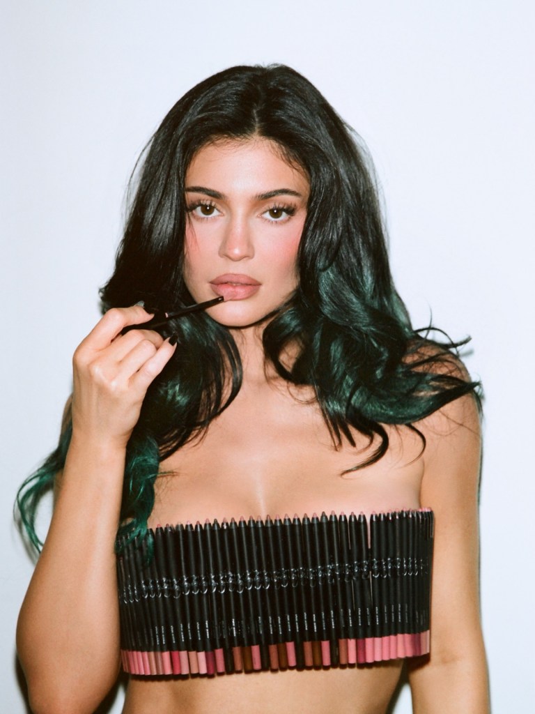 Kylie Jenner wearing a strapless top made of lip liner pencils, holding another lip pencil to her mouth.
