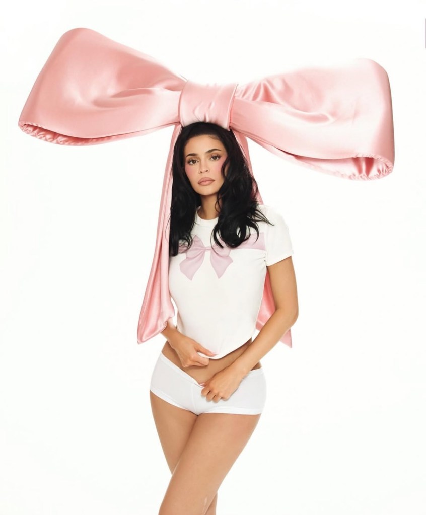 Kylie Jenner wearing a giant pink bow headpiece, a white crop top with a pink bow, and white underwear.