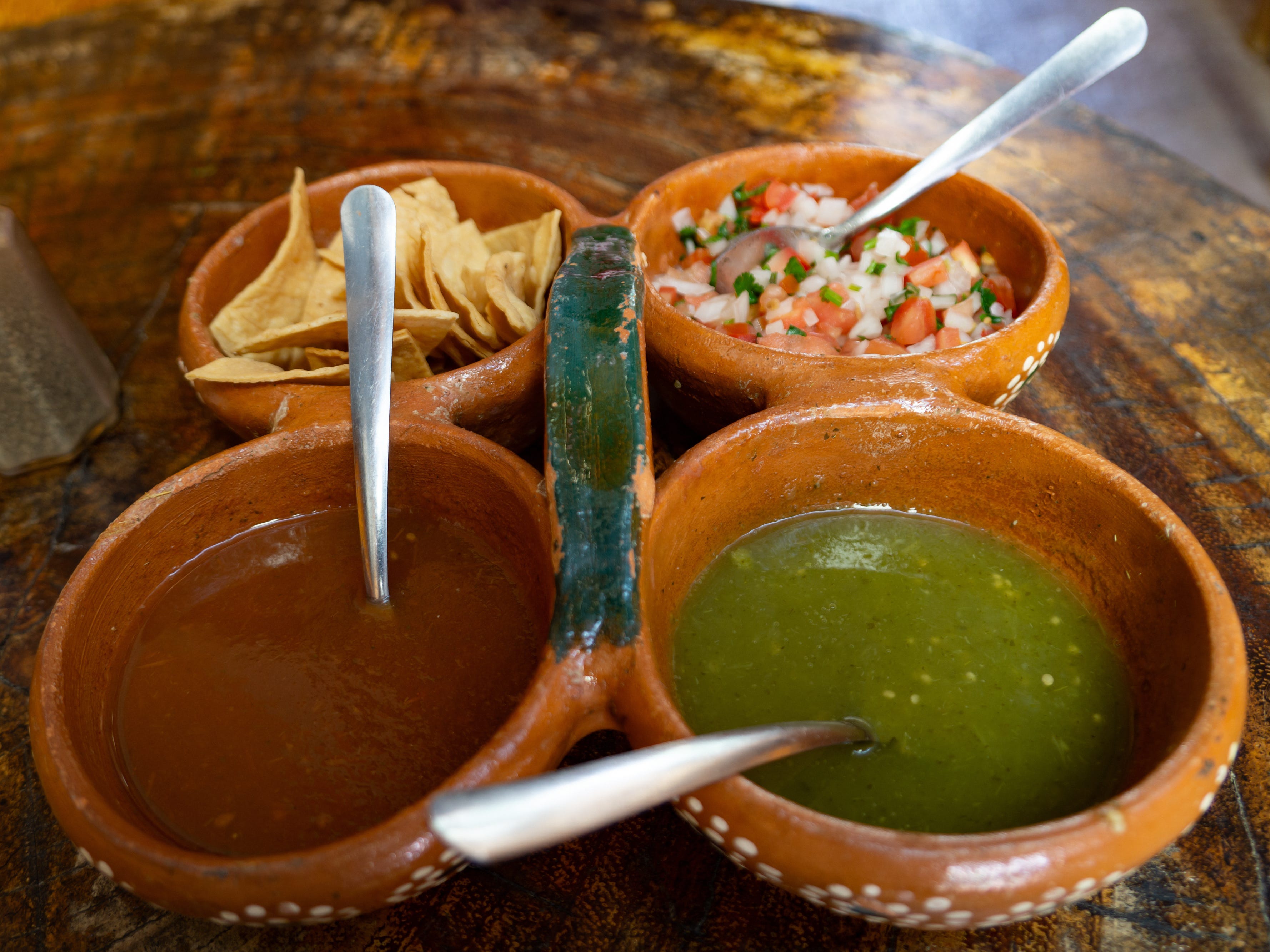 Small ceramic bowls with red and green salsas, pico de gallo, and chips on a table at a Mexican restaurant.
