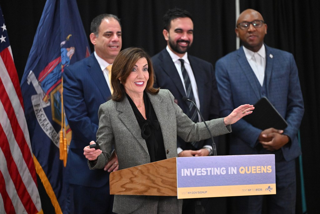 Kathy Hochul speaking at a podium with “Investing in Queens” on the front, flanked by three men, with American and New York flags in the background.