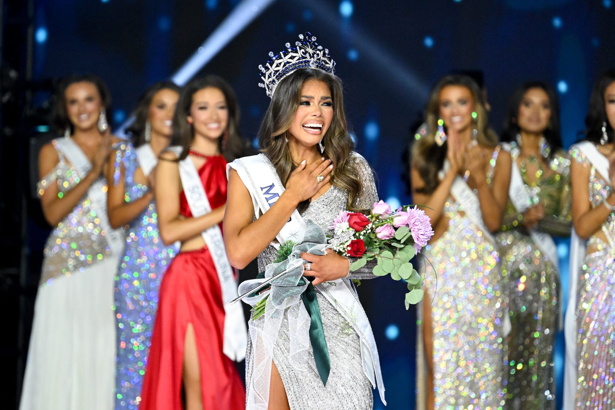 Alma Cooper holding flowers and wearing a crown at Miss USA 2024, with other women standing behind her.