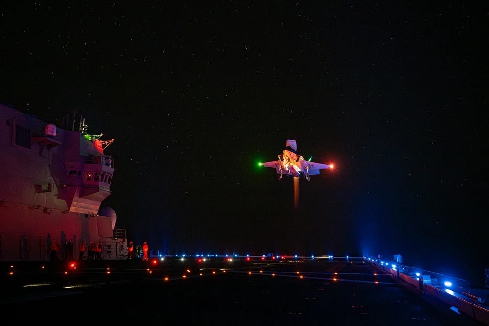 An F-35B lands on the illuminated flight deck of the JS Kaga during a night flight test.