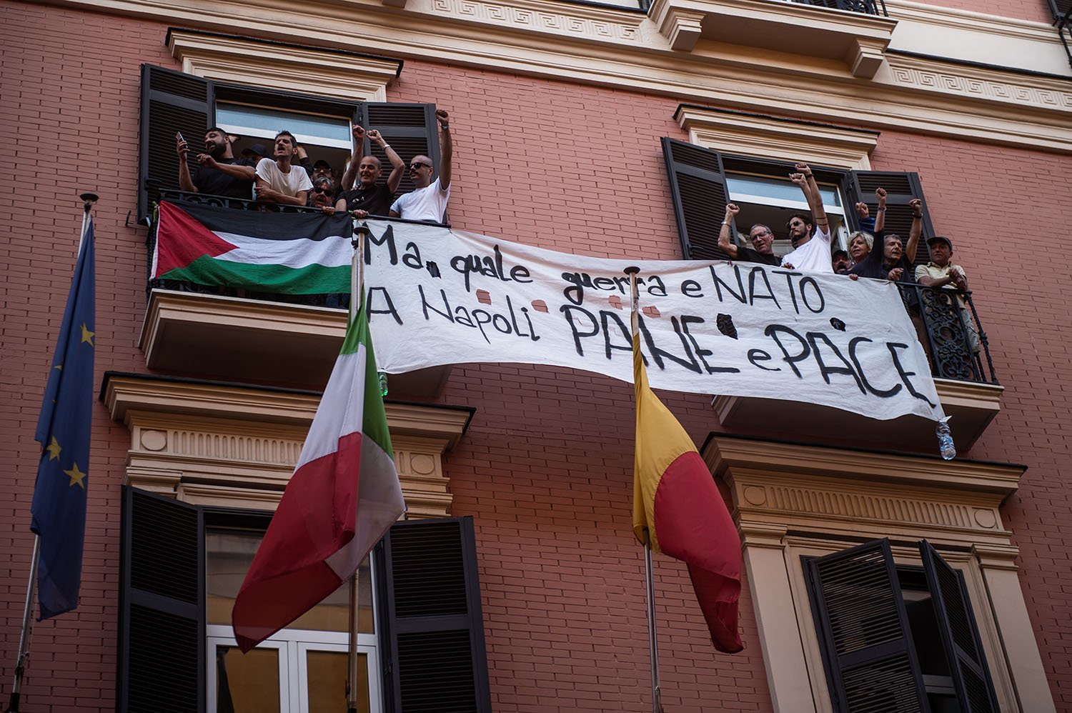 A large white banner with Neopolitan words on it stretches between two balconies where people lean out and raise their fists. The flags of the EU, Italy, and Naples fly in front of the building.