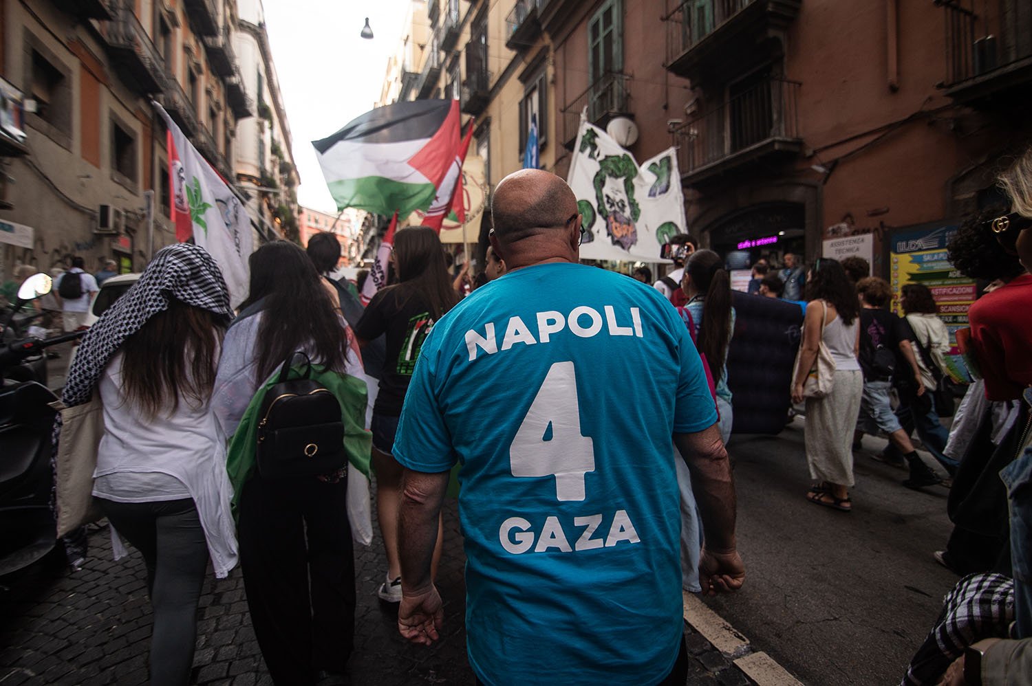 A man walks behind a group of protesters holding Palestinian flags down a street with buildings on either side.