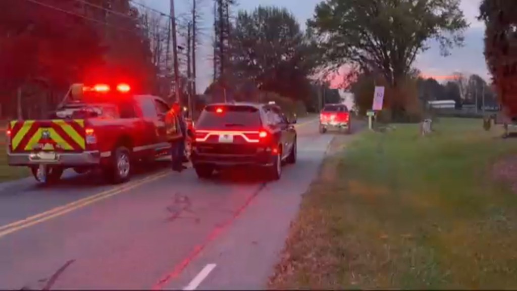 A red utility truck with flashing red lights and a person standing next to a black SUV on a rural road.