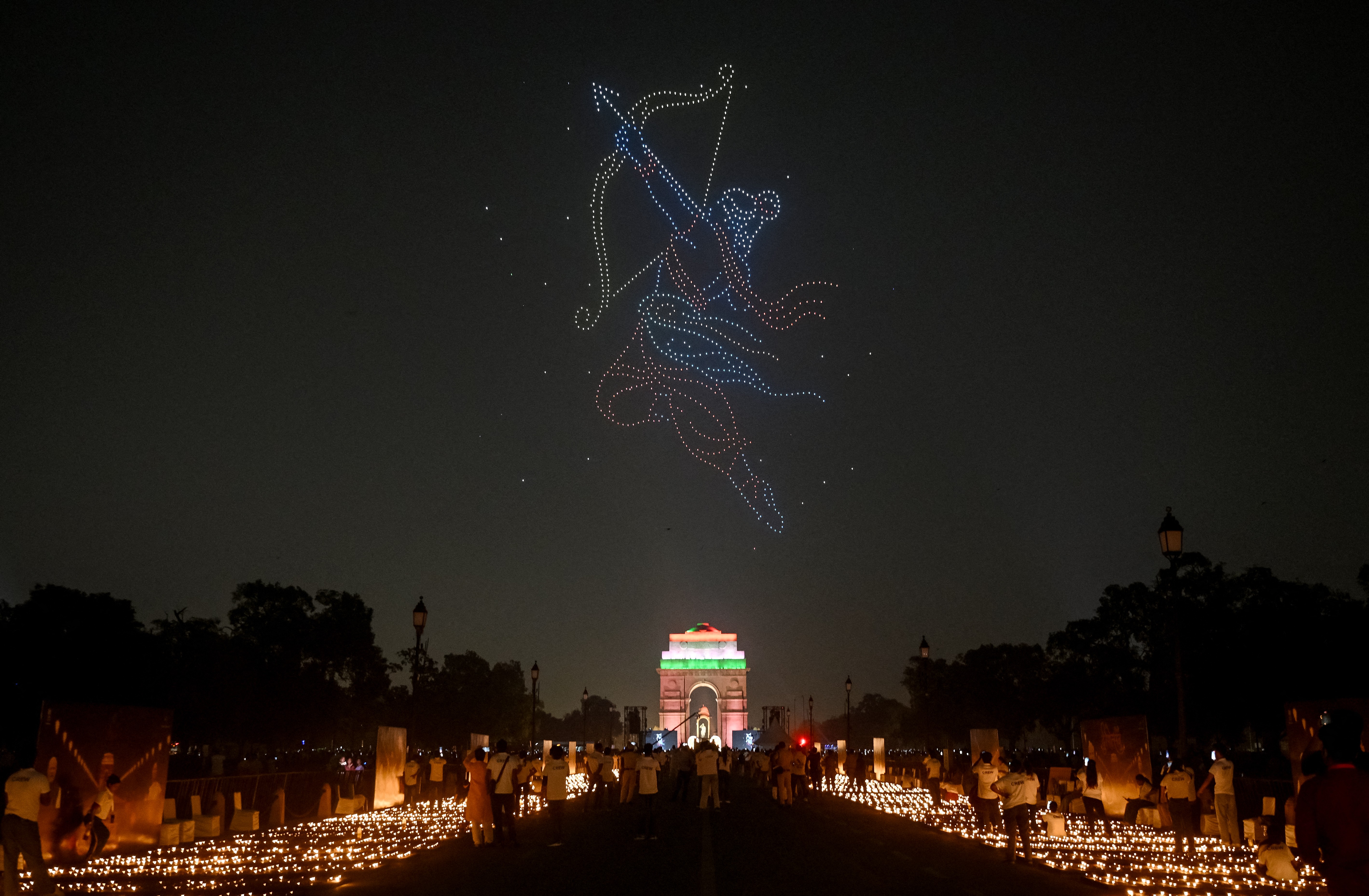 Thousands of lit oil lamps, arranged on the ground beneath a drone light show depicting a mythical Hindu character with a bow and arrow.