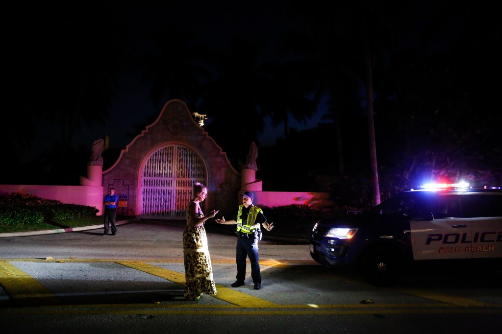 Woman talks to a Palm Beach police officer in front of Mar-a-Lago at night, with a police car nearby.