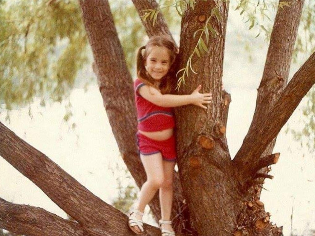 Little girl in red outfit holding onto a tree.