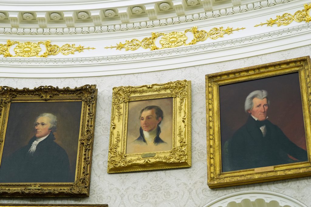 Portraits of Alexander Hamilton, James Monroe, and Andrew Jackson hanging on a curved wall with ornate gold and white trim.