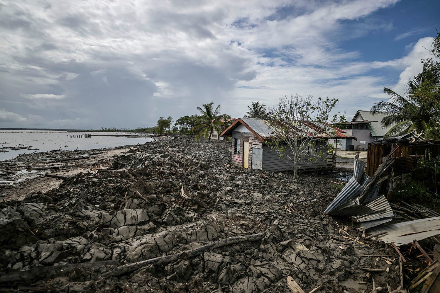 A small house with a corrugated metal roof sits on a craggy, broken up coastline with debris around it against a partly cloudy sky.